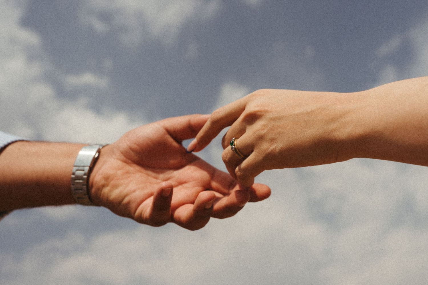 Close-up of two hands reaching towards each other against a cloudy sky.