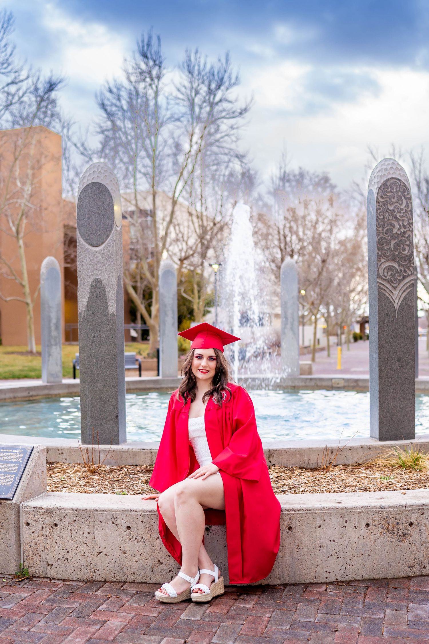 Graduate in red cap and gown poses by a fountain on campus during graduation photos.