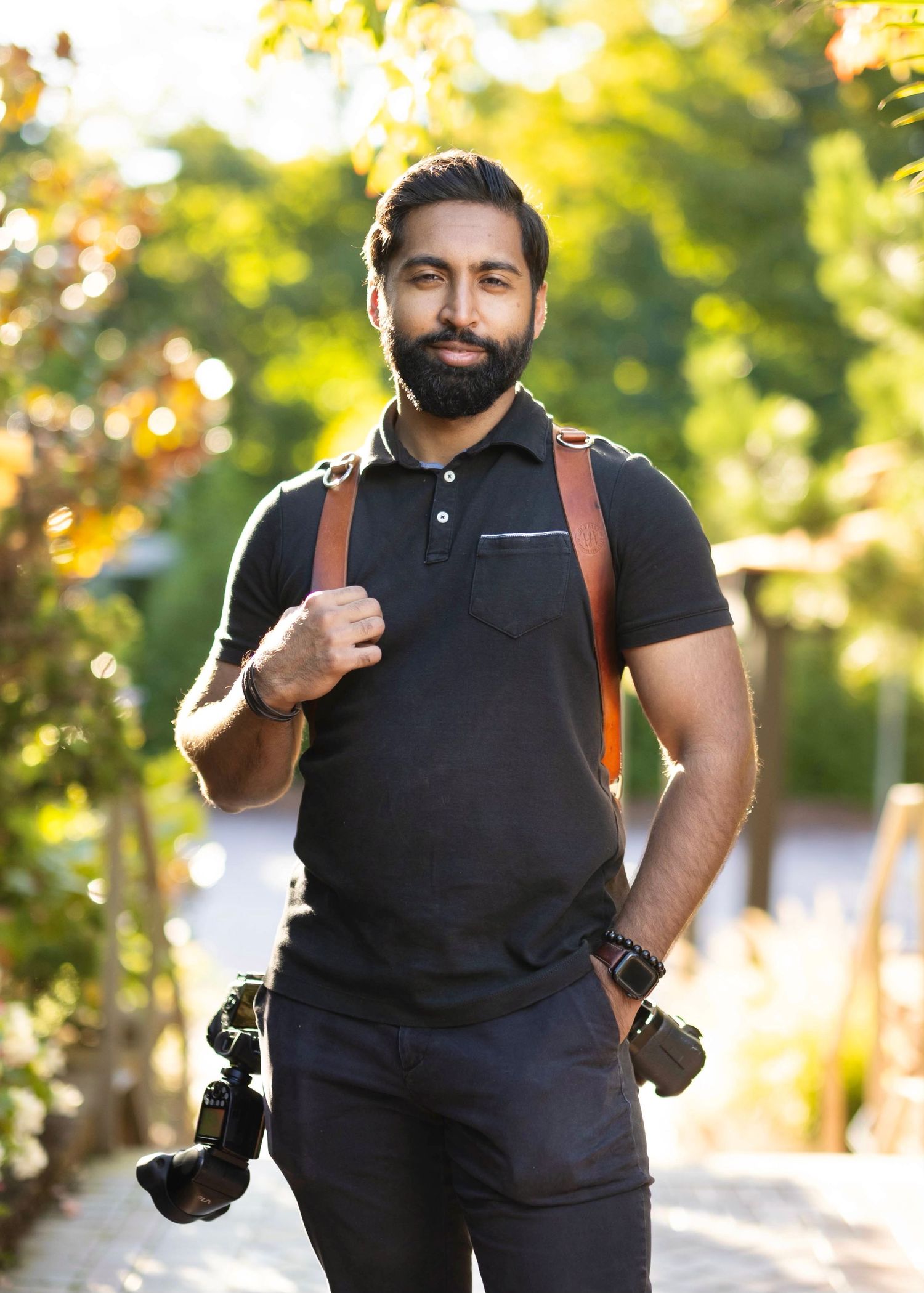 A professional photographer in black attire with leather camera straps poses outdoors in natural lighting.
