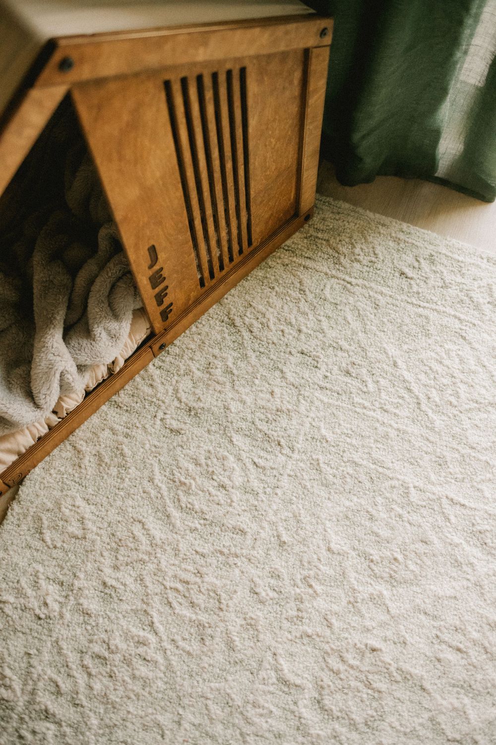 White fluffy area rug beside wooden dog crate with folded blanket inside near green curtain.