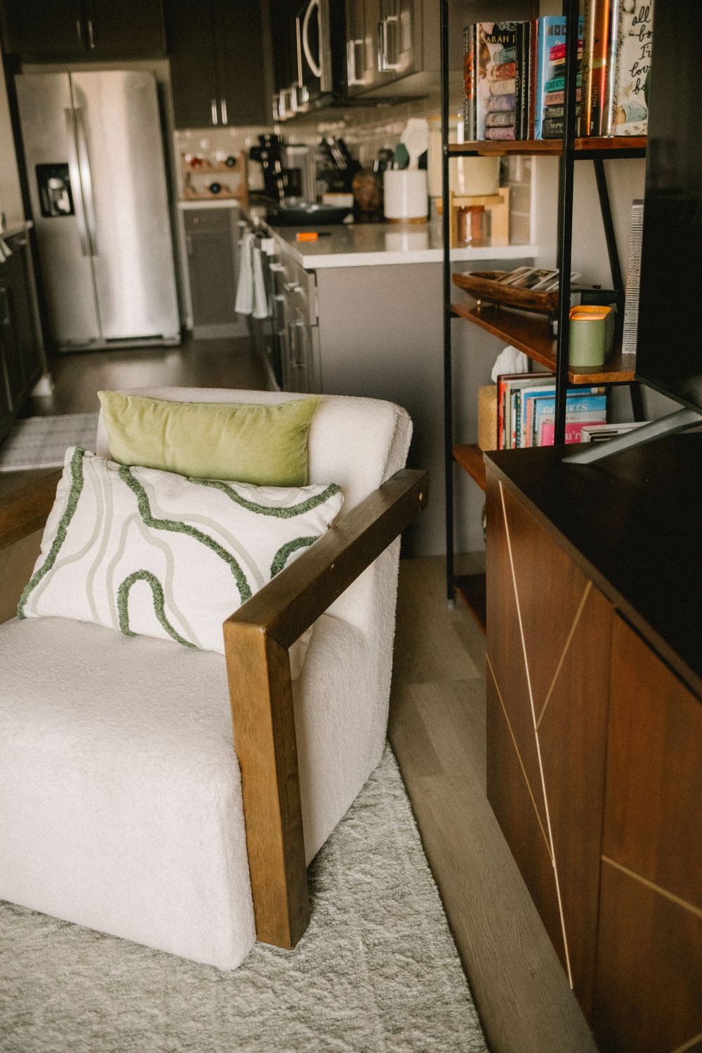 White boucle armchair with wood frame and green pillow beside bookshelf in modern apartment.