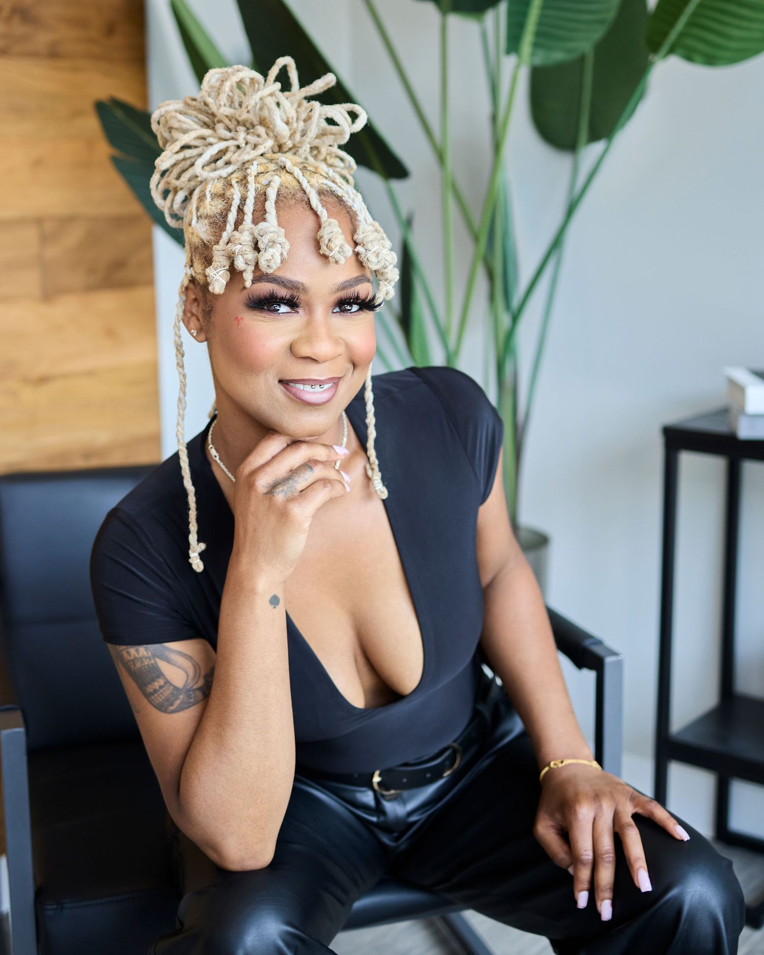 Woman with blonde braided hairstyle in black top posing by indoor plant.