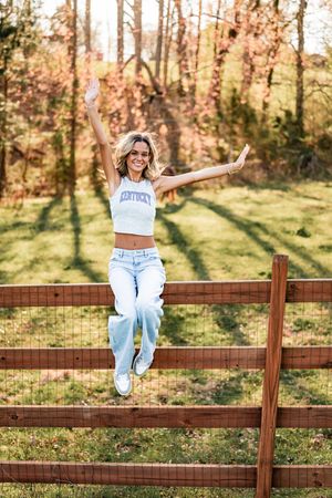 A person in light wash jeans and white crop top sits joyfully on a wooden fence in a sunny autumn setting.