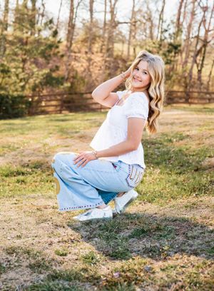 High School Senior in white t-shirt and light blue jeans crouches outdoors in a sunny wooded setting wearing sneakers.