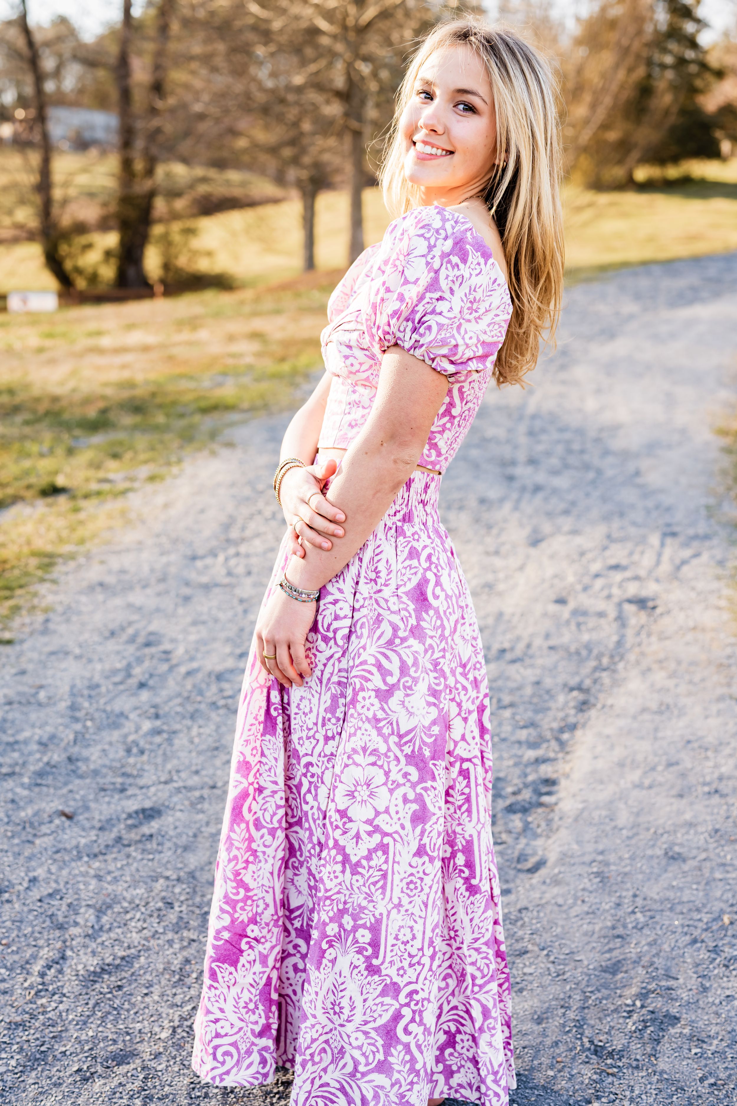A High School Senior wearing a pink and white floral maxi dress poses on a gravel path surrounded by autumn trees.