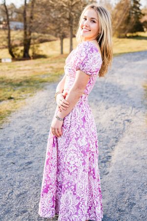 A High School Senior wearing a pink and white floral maxi dress poses on a gravel path surrounded by autumn trees.