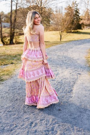 High School Senior wearing a pink boho style two-piece outfit with a flowing tiered skirt poses on a gravel path during golden hour.