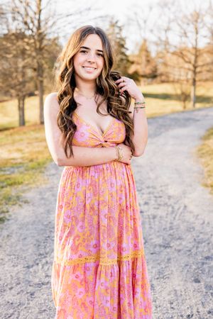 High School Senior wearing a pink floral maxi dress with long wavy hair stands on a gravel path during golden hour.