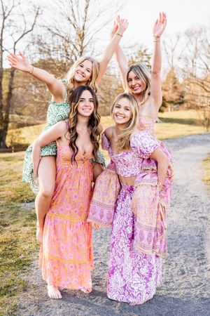 High School Seniors in colorful floral maxi dresses pose joyfully with raised arms on a path during golden hour.