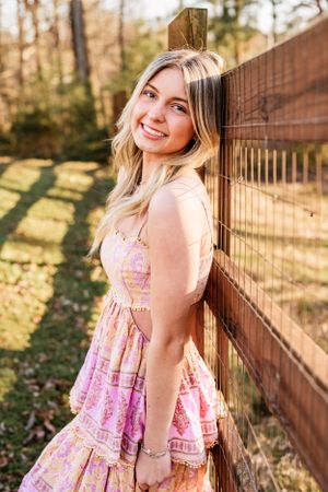 A High School Senior in a pink patterned dress leans against a wooden fence on a sunny autumn day, casting long shadows.