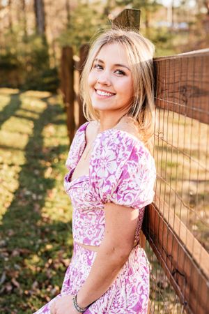 A High School Senior in a pink and white floral dress leans against a wooden fence on a sunny day in a backyard setting.