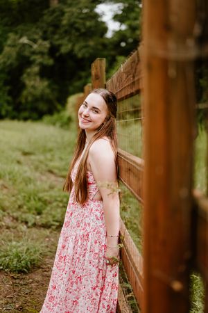 Someone in a floral pink summer dress poses casually against a rustic wooden fence in a natural outdoor setting.