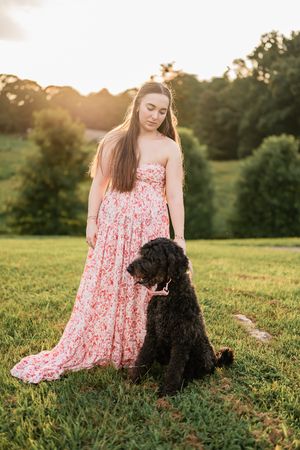 A sunset photoshoot in a field featuring someone in a pink dress sitting with their black dog.