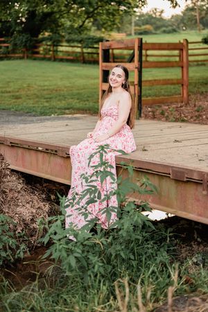 A whimsical maternity photo series shows a floral pink dress against a rustic wooden bridge in a garden setting.