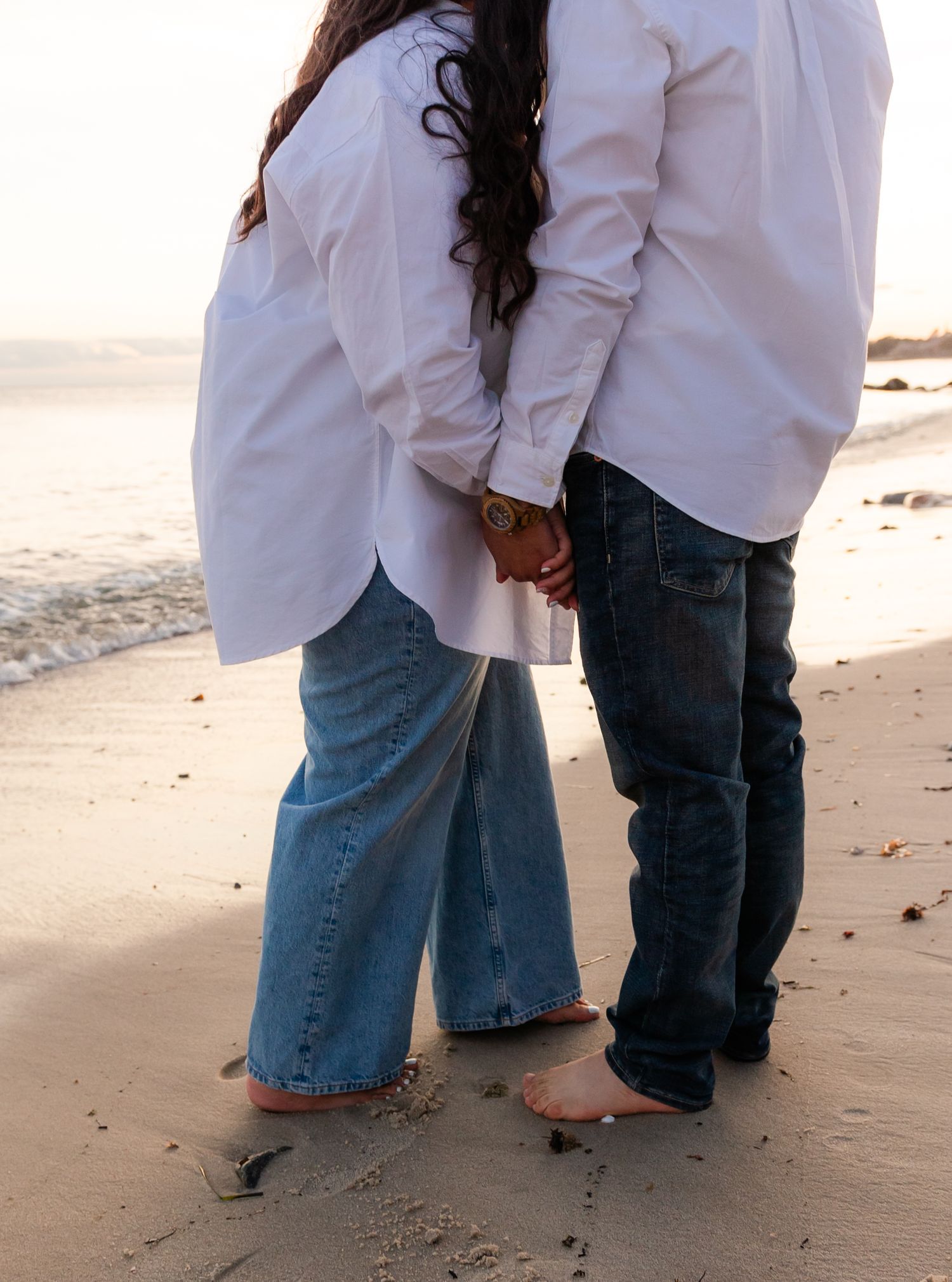 Two people standing close together on a beach, wearing white shirts and jeans.