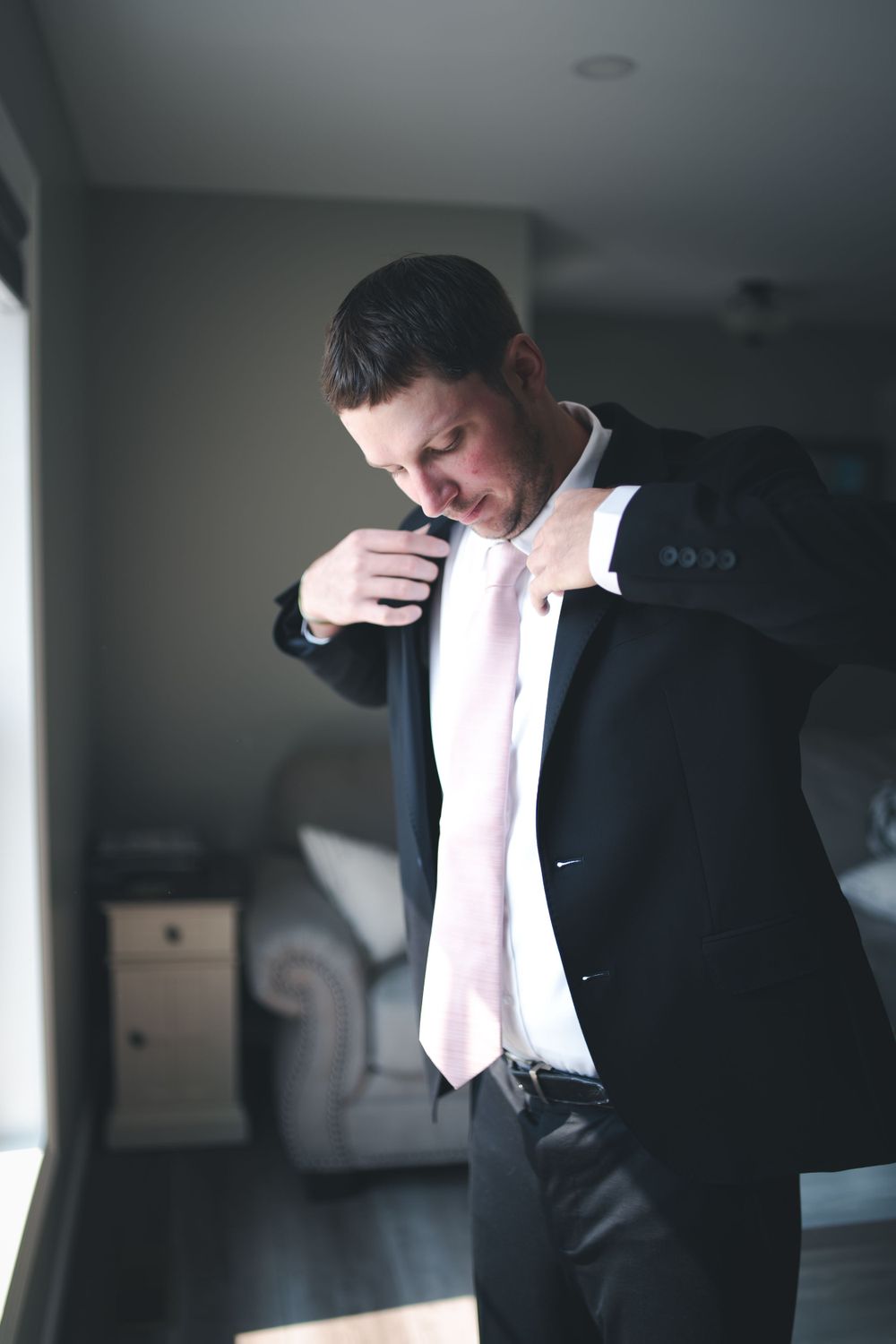 A groom adjusts his tie while getting ready in formal attire near a window.