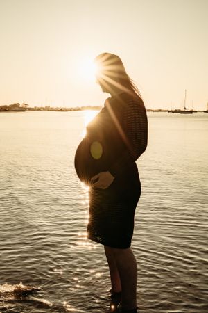 Silhouette of an expectant mother standing by the water during golden hour sunset.