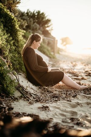Person in brown sweater sitting on beach during sunset with greenery backdrop.