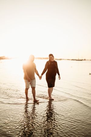 Couple holding hands while walking in shallow beach water during sunset.