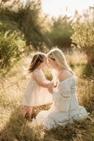 Intimate mother-daughter moment in sunlit field wearing flowing white dresses.
