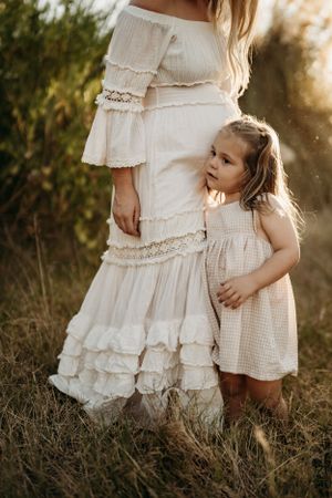 Child in white dress standing close to mother's flowing white dress in golden sunlight.
