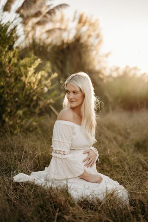 Woman in white off-shoulder dress sits gracefully in a sunlit field with tall grass and golden sunset glow.