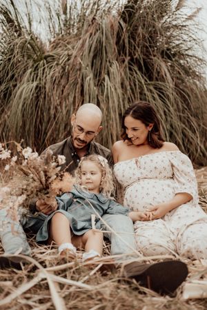 Family portrait with expecting mother in white dress and child in blue dress sitting together in front of pampas grass.