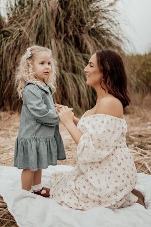 Mother in floral dress holds hands with daughter in denim dress against natural backdrop.