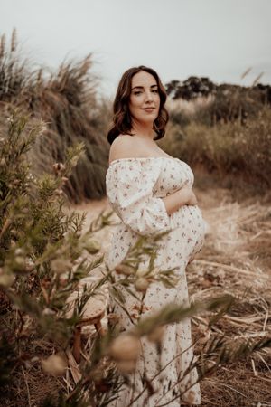 Maternity portrait of woman in white floral dress posing in natural outdoor setting.
