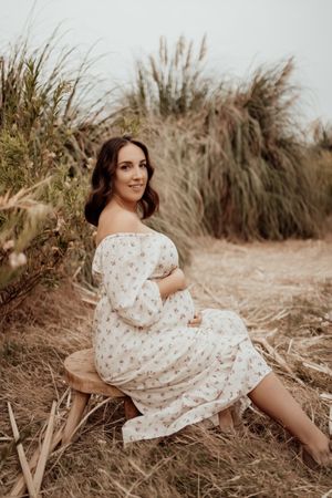 Expectant mother in floral dress sitting gracefully among dried grass and foliage.