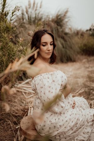 Side profile of pregnant woman in white dress posed among wild grasses.