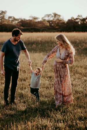 A family holds hands in a rustic field during golden hour while their toddler jumps with joy.