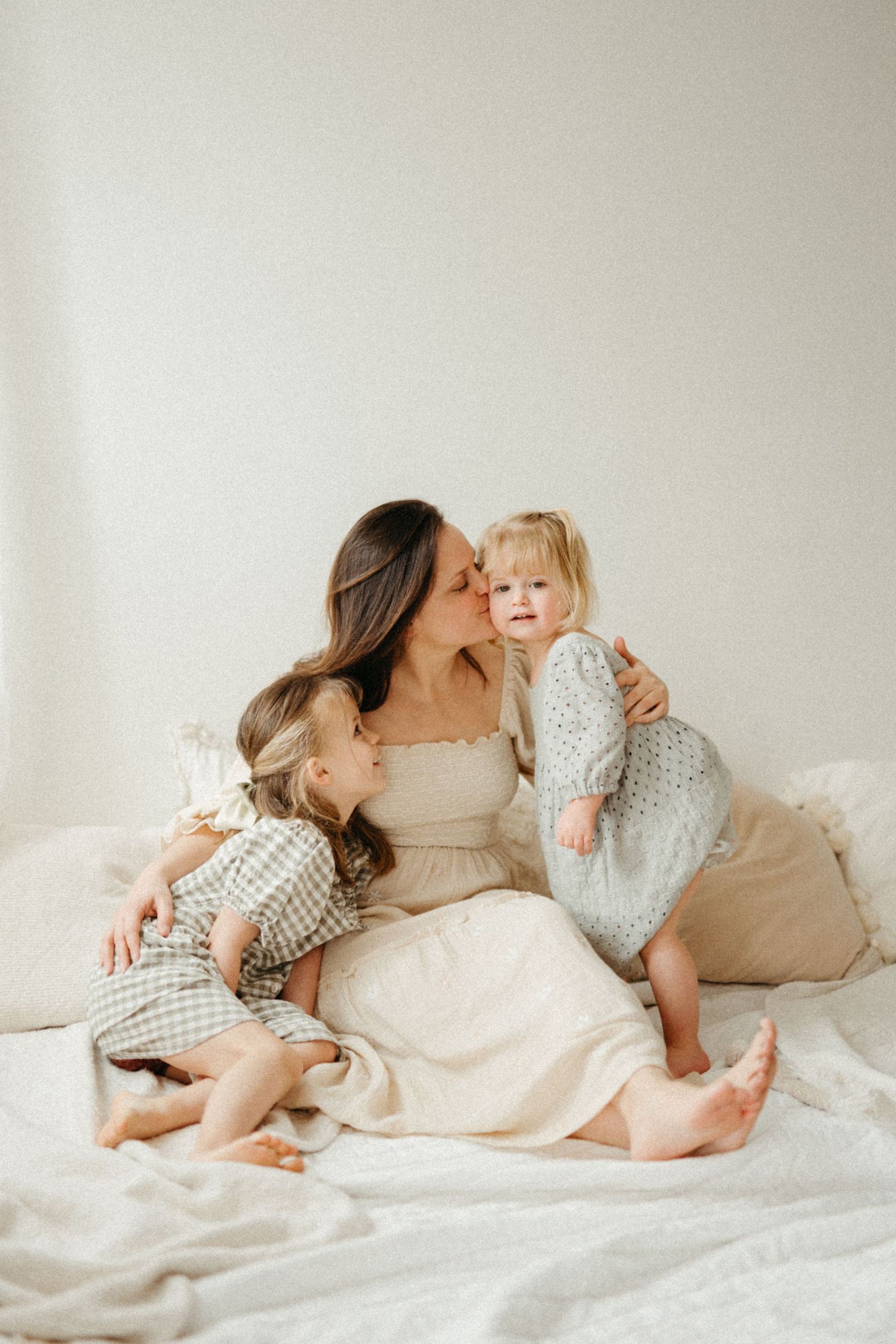 A mother sits on a white bed kissing her toddler daughter while another young child leans against her.