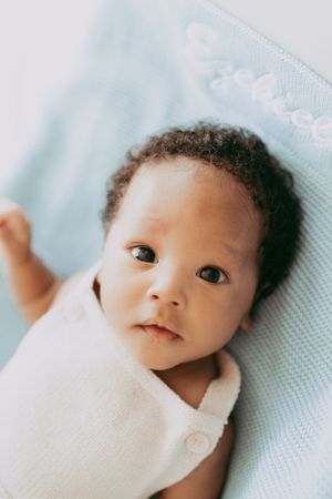 Sweet newborn baby wrapped in white swaddle on light blue blanket looking at camera.