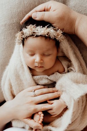 Sleeping newborn wearing delicate floral crown wrapped in soft beige blanket.