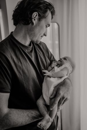 Intimate black and white photo of father holding tiny newborn against window light.