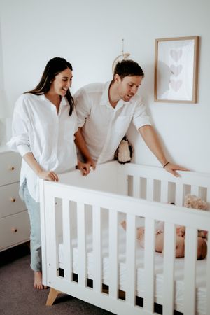 Parents leaning over white crib together in bright modern nursery setting.