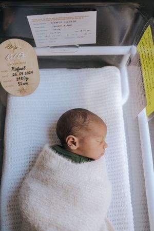 Newborn baby sleeping peacefully swaddled in white blanket in hospital bassinet.