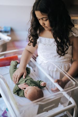 Young child in white lace dress tenderly observing baby in hospital bassinet.