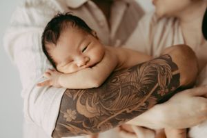 A sleeping newborn rests peacefully on a tattooed arm wearing a white outfit.