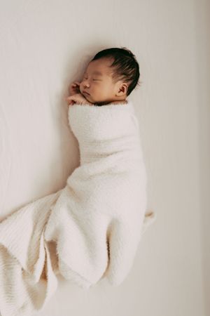 A sleeping newborn baby wrapped snugly in a soft white blanket against a light background.