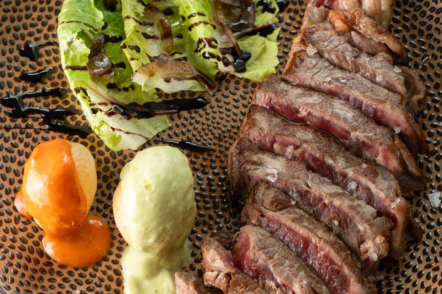 Grilled sliced steak served with fresh lettuce and citrus on a dark textured surface.