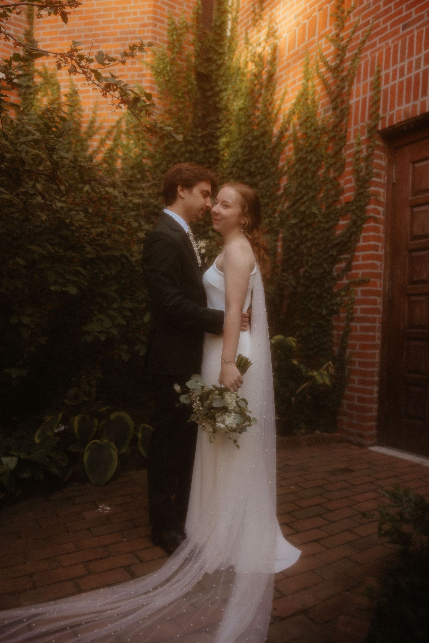 Romantic wedding couple embracing against brick wall with greenery at sunset.