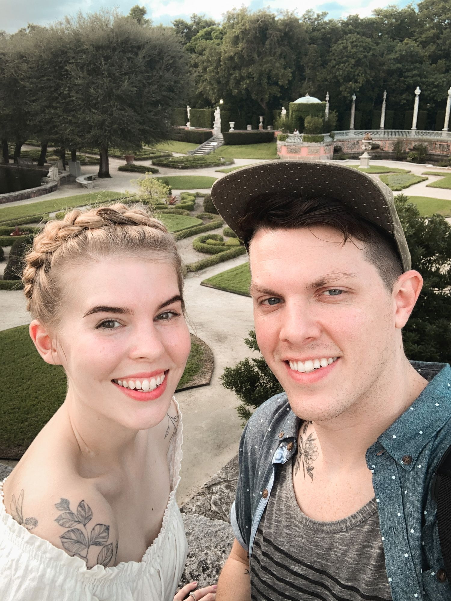 A couple takes a selfie overlooking ornate gardens with decorative pathways and sculptures.