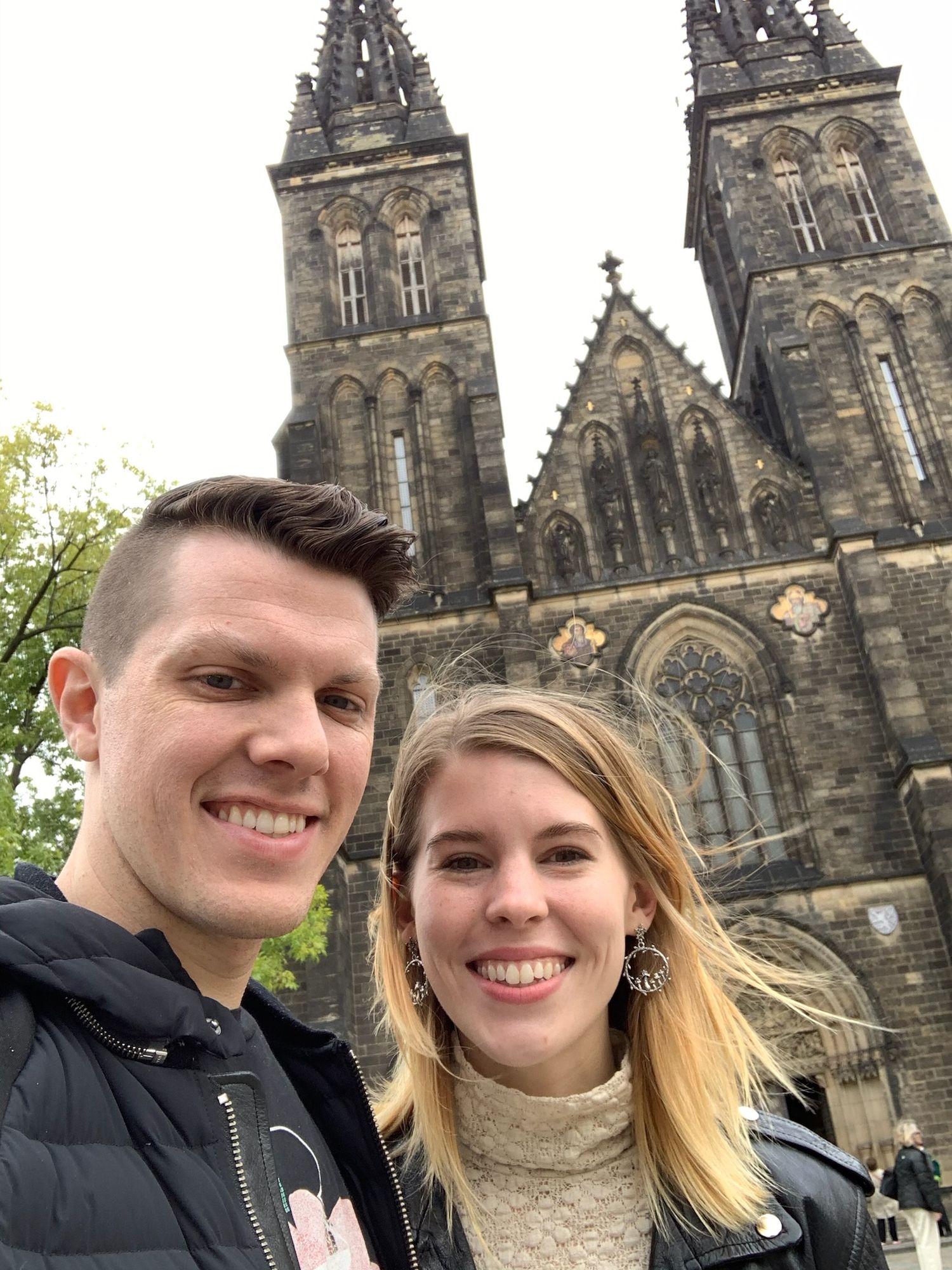A smiling couple takes a selfie in front of a grand gothic cathedral with stone spires.