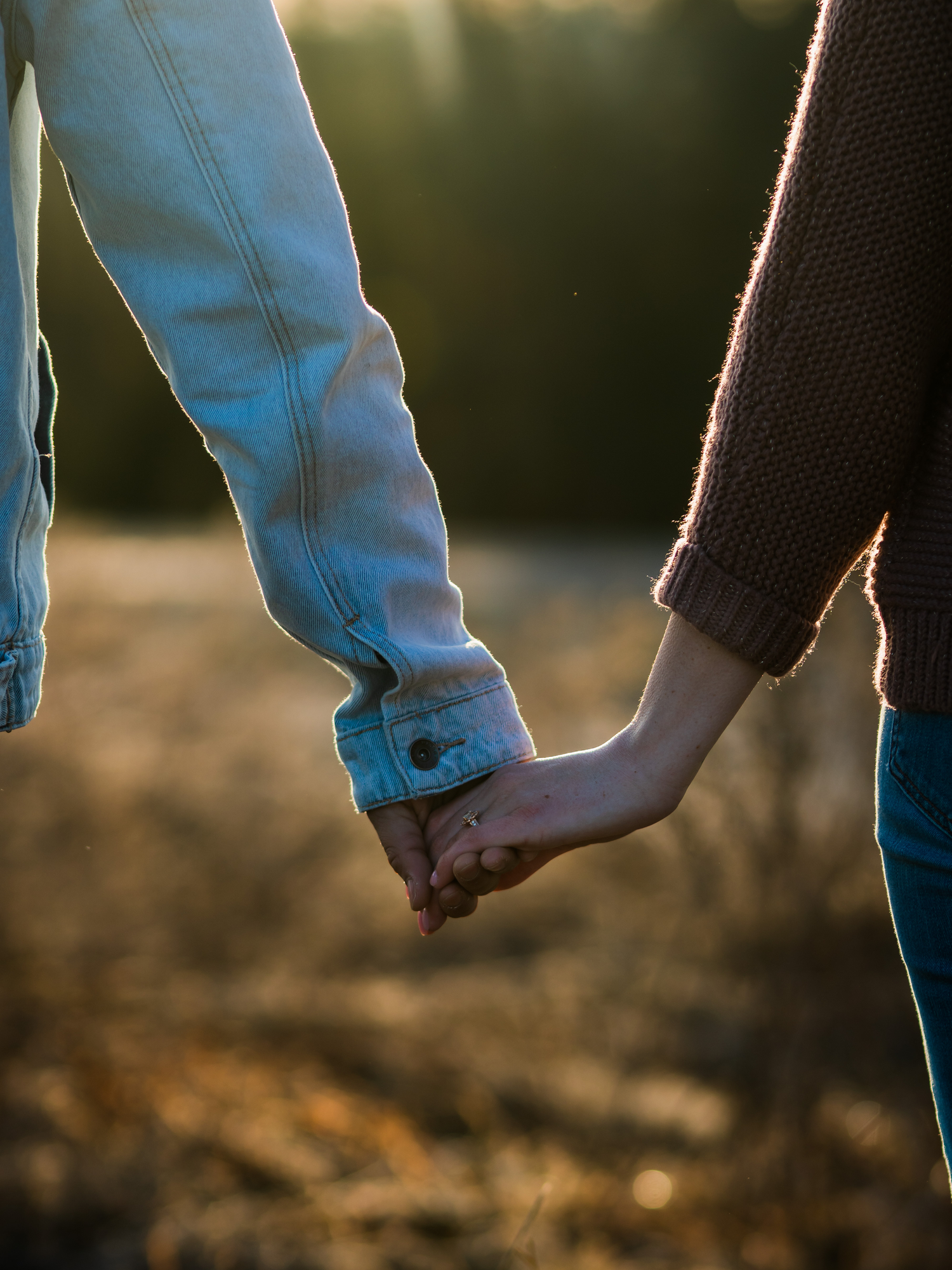 Close up of two people holding hands at sunset in a field with golden light filtering through.
