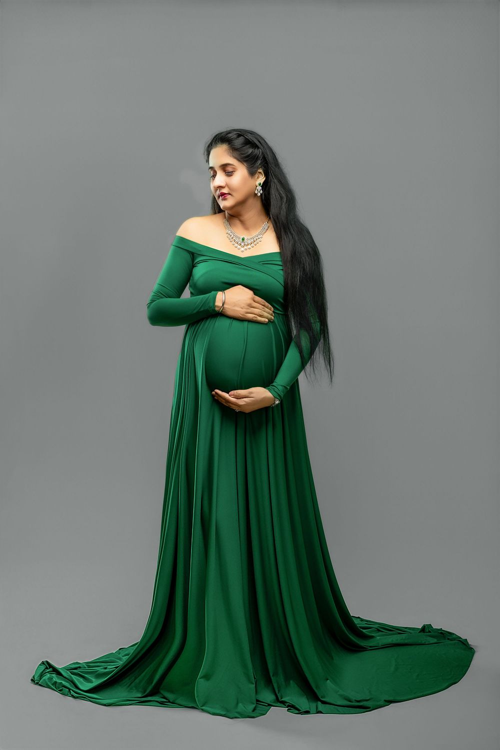 A woman in a flowing emerald green maternity gown poses gracefully against a gray studio backdrop.