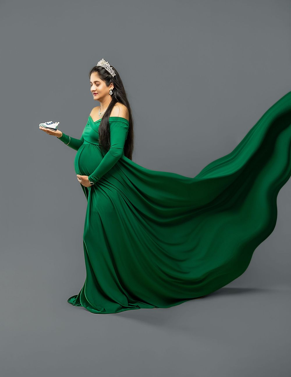 A pregnant woman in a flowing emerald green gown and tiara poses elegantly against a gray backdrop.