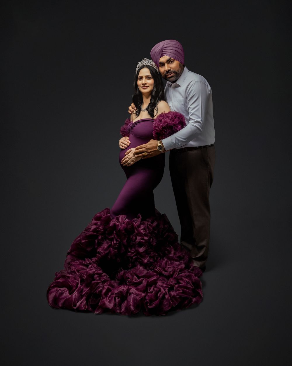 Couple in elegant attire pose intimately against dark backdrop, with woman wearing dramatic purple ruffled gown.
