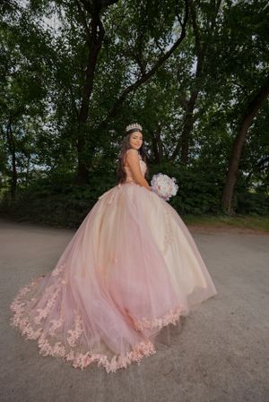 Person in blush pink ball gown with floral details poses on path surrounded by trees.